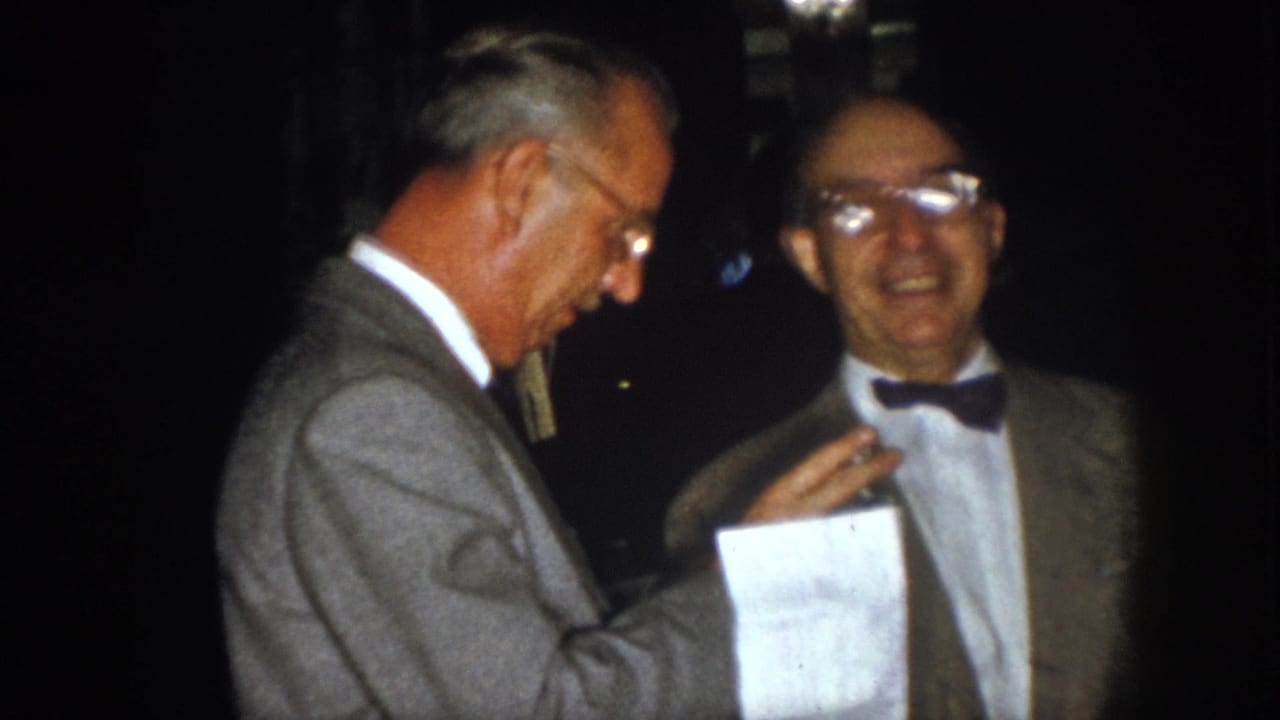 1958 Virginia home-movie frame of two older men in formal jackets at night, with one holding a paper during conversation.