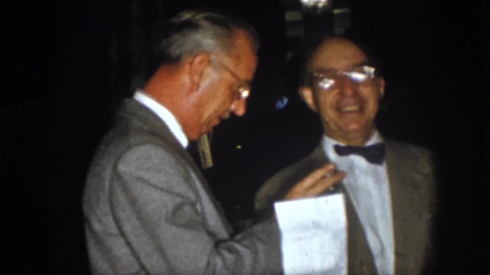 1958 Virginia home-movie frame of two older men in formal jackets at night, with one holding a paper during conversation.