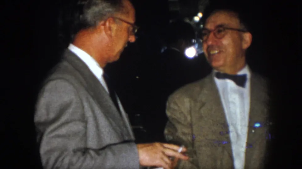 1958 Virginia home-movie frame of two older men smiling in conversation, one wearing a bow tie and light jacket.