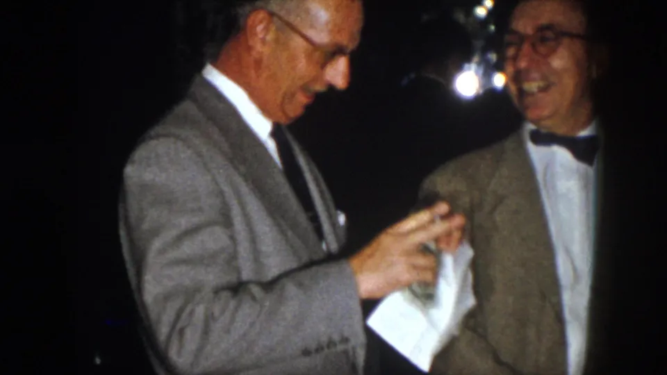 1958 Virginia frame of two formally dressed older men at night, with one holding a paper as they converse.