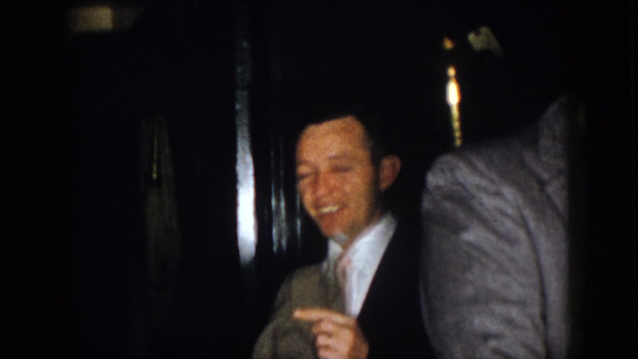 1958 Virginia 8mm frame showing two suited adult men in a dim interior, with one smiling broadly while gesturing toward the camera.