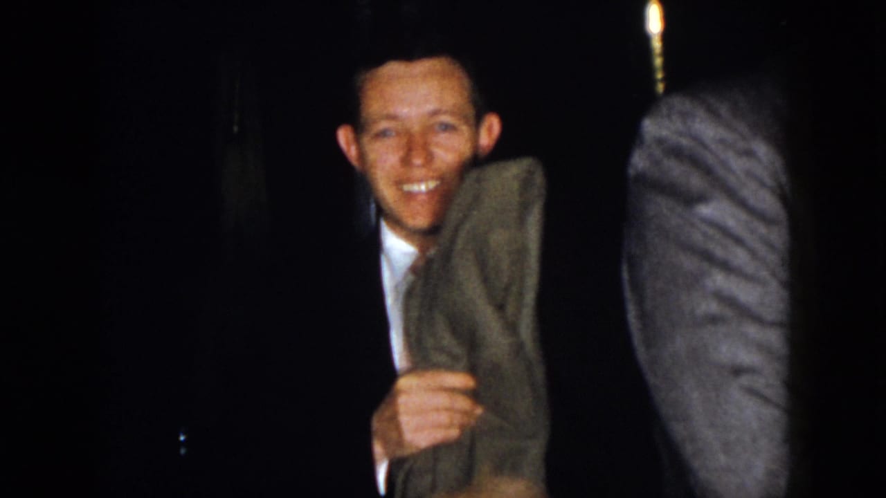 A smiling man in a dark suit holds a coat while another suited figure is partly visible nearby.