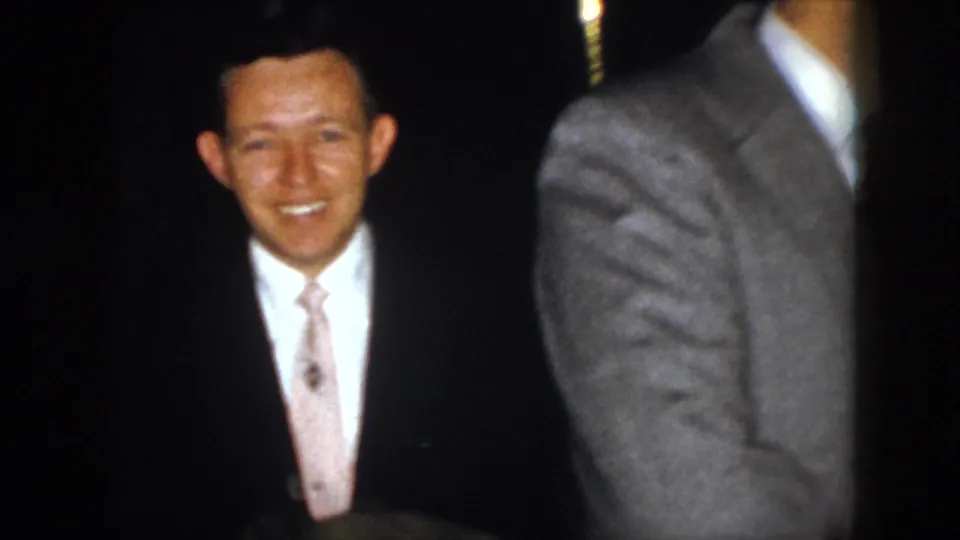 1958 Virginia 8mm frame showing a camera-facing man in suit and tie, partly blocked by another suited adult in the foreground.