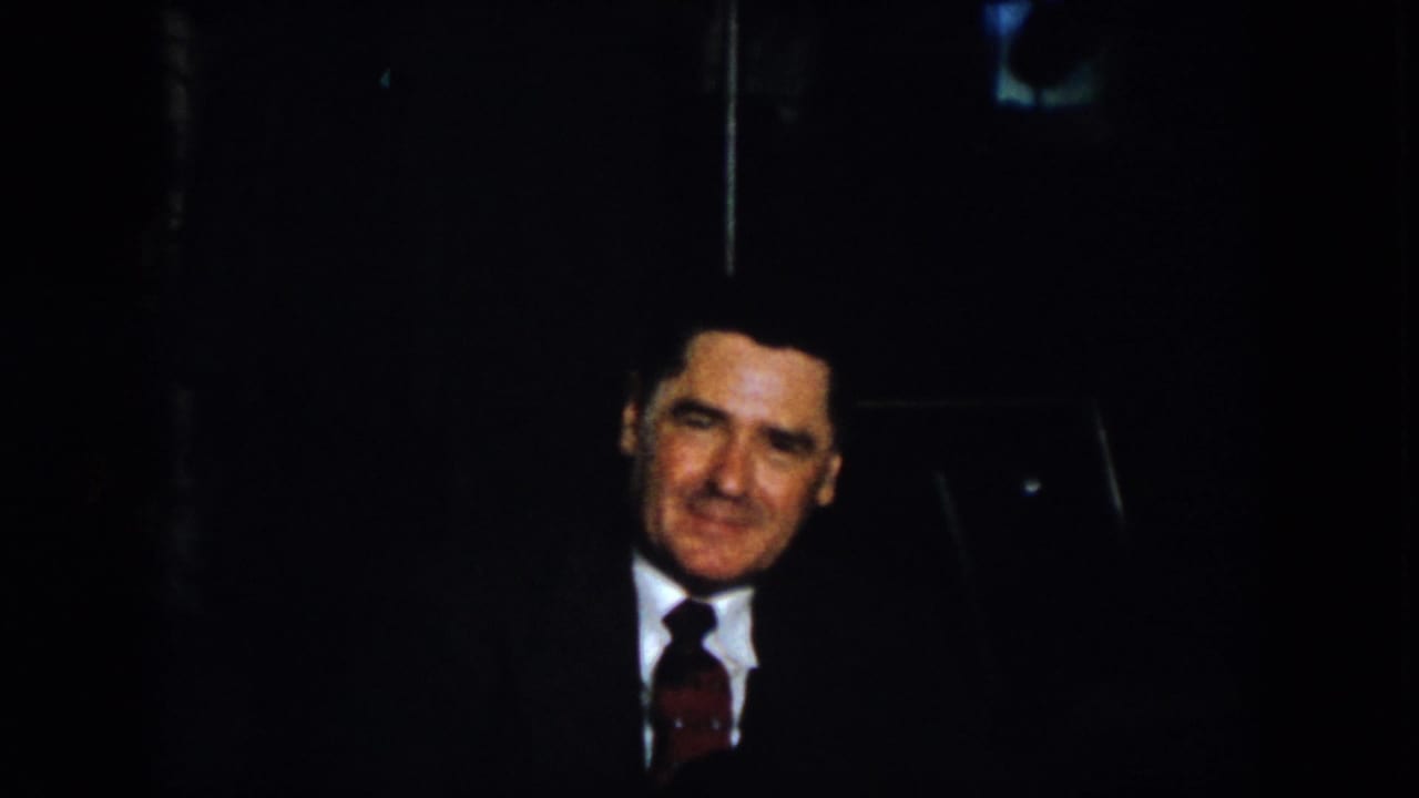 1958 Virginia 8mm frame showing a suited man with a tie facing the camera in a dark indoor setting.