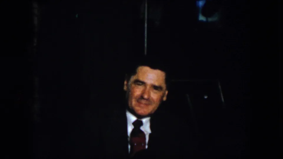 1958 Virginia 8mm frame showing a suited man with a tie facing the camera in a dark indoor setting.