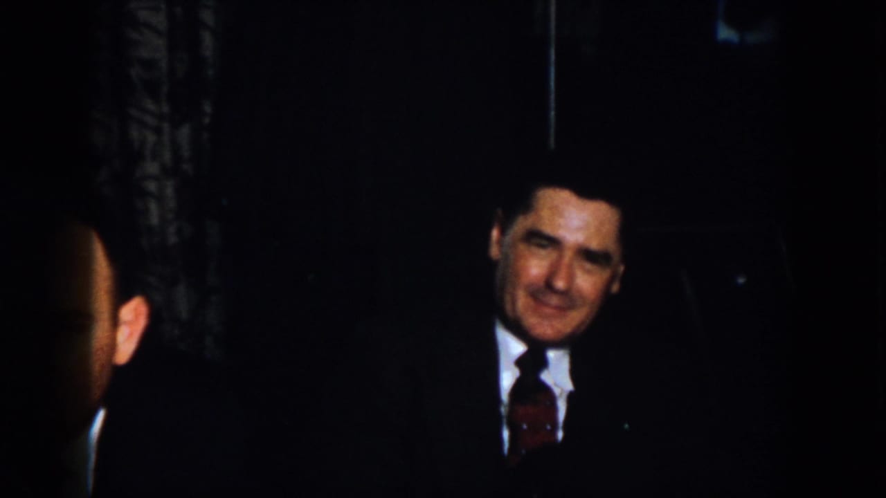 1958 Virginia home-movie frame with a suited man smiling in a shadowy interior, partially cropped by the contact sheet.