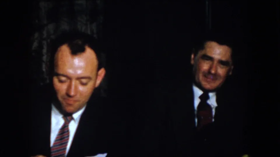 1958 Virginia 8mm frame showing two suited men indoors, one looking down and another watching from the center.