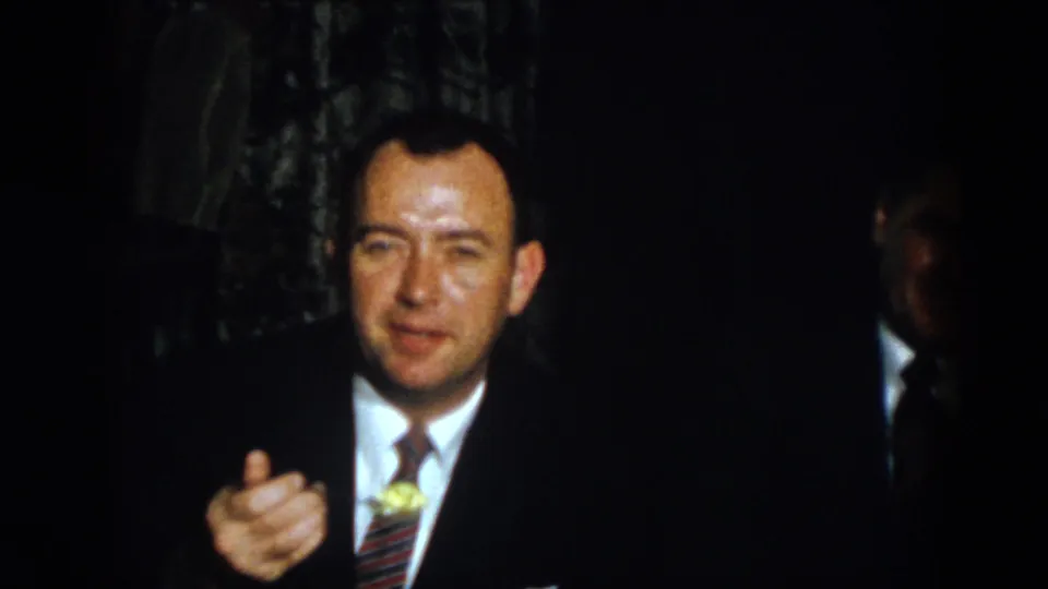 1958 Virginia home-movie frame of a suited man gesturing as he speaks, with another cropped figure at the edge.