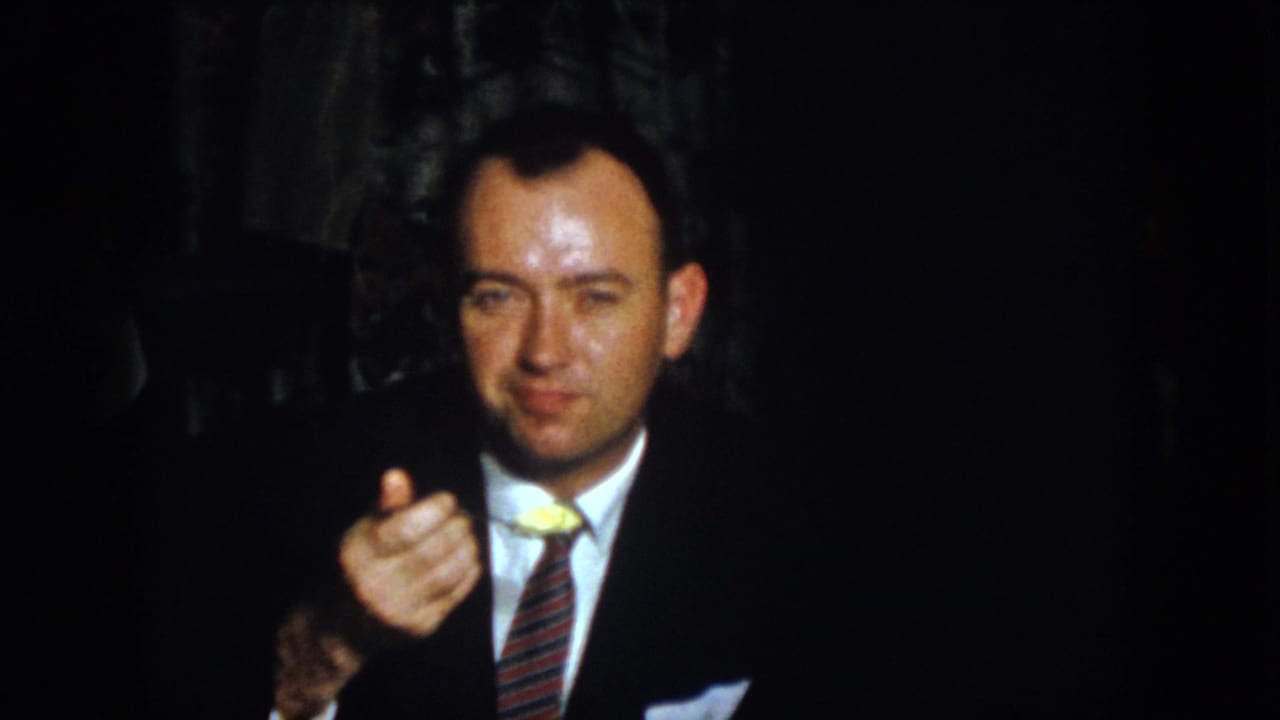 Suited man holds a spoon with a small yellow food item in a dim interior.