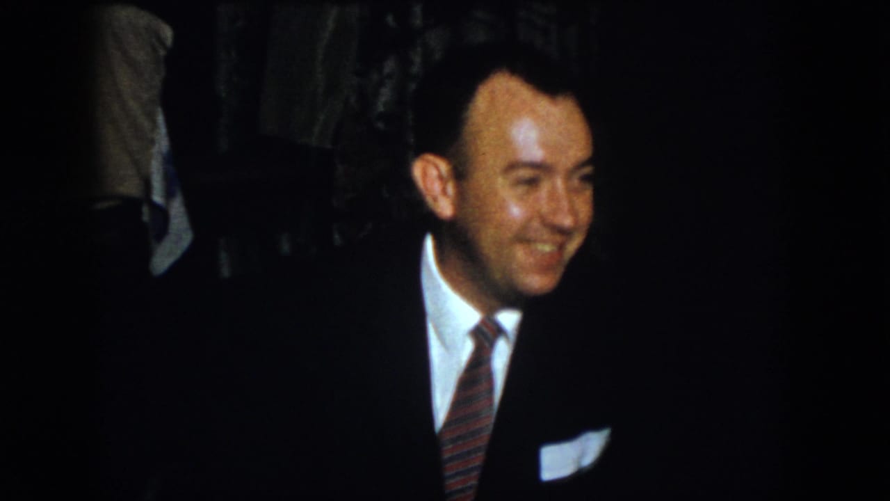 Suited man smiles while looking off to the side in a dark room.
