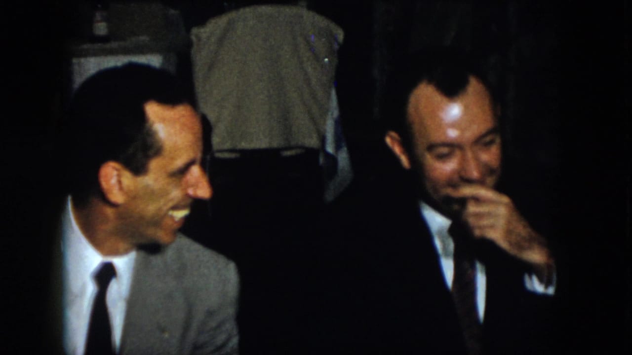 Two adult men, one in a light suit and one in a dark suit, laugh together during a candid 1958 Virginia home-movie moment.