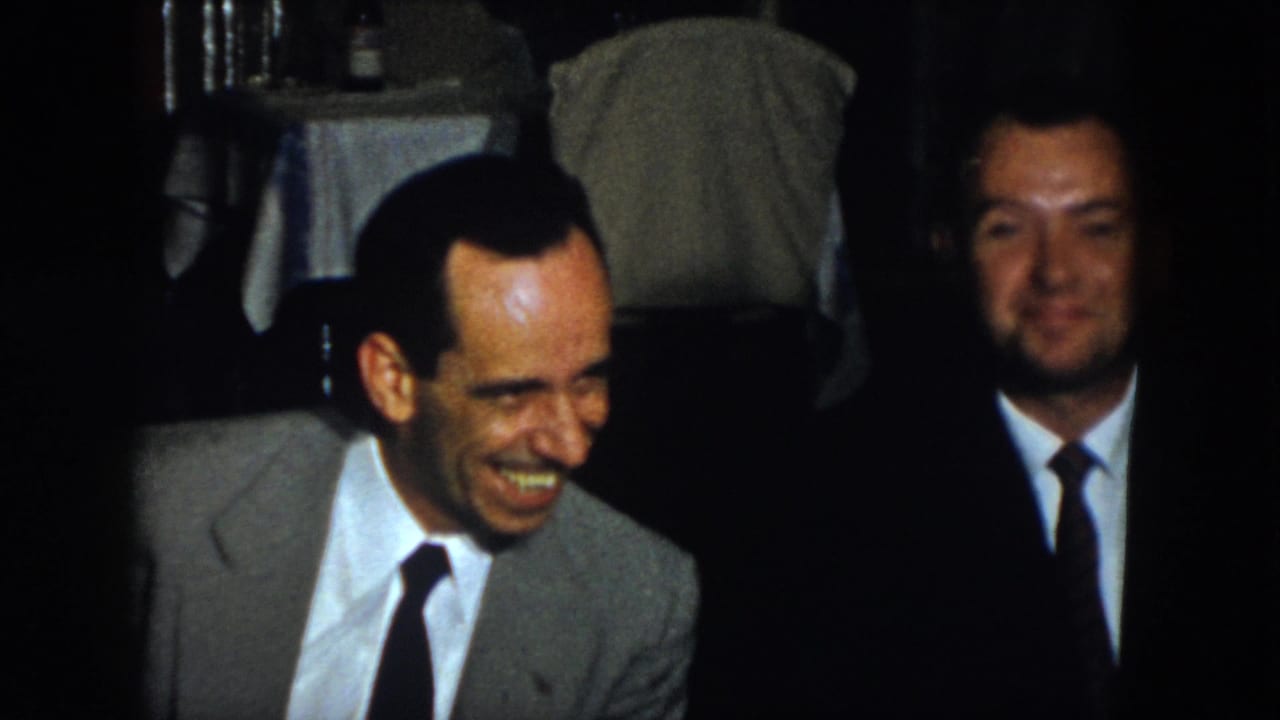 Two suited men sit in a dim interior, with the man at left laughing broadly.