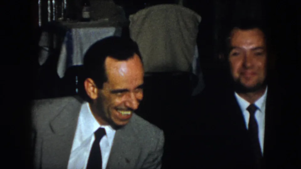 In 1958 Virginia home-movie footage, two adult men in suits sit in a dim restaurant or private dining room as the man at left laughs and the man at center faces forward.