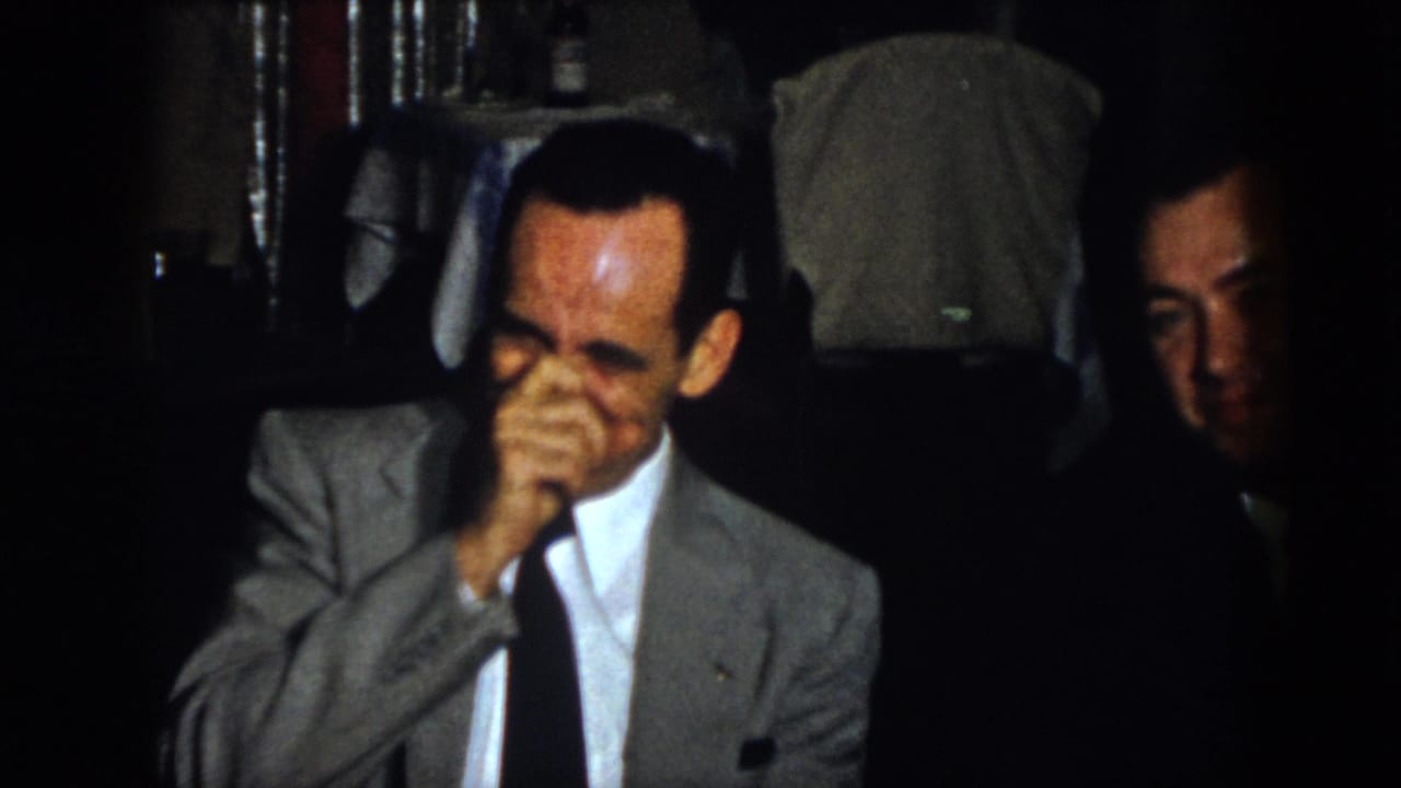 In the same 1958 Virginia interior, a man in a gray suit covers his mouth while laughing, with another adult man partly visible at the right edge.