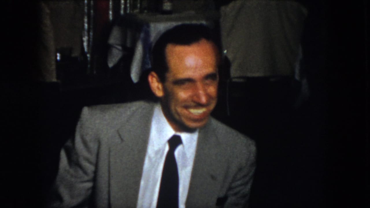 The 1958 Virginia home-movie frame shows an adult man in a gray suit smiling in low light, with a mostly obscured companion and dining-room details behind him.