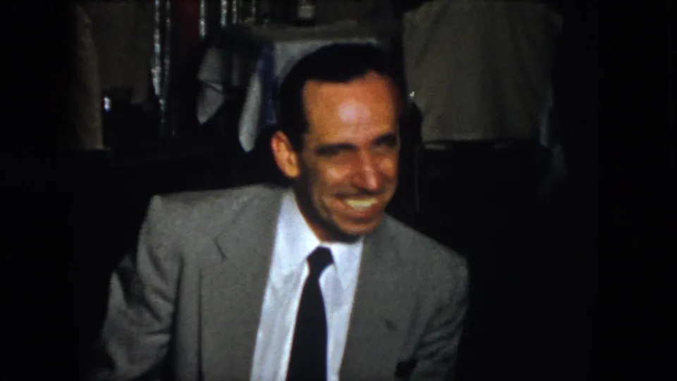 The 1958 Virginia home-movie frame shows an adult man in a gray suit smiling in low light, with a mostly obscured companion and dining-room details behind him.