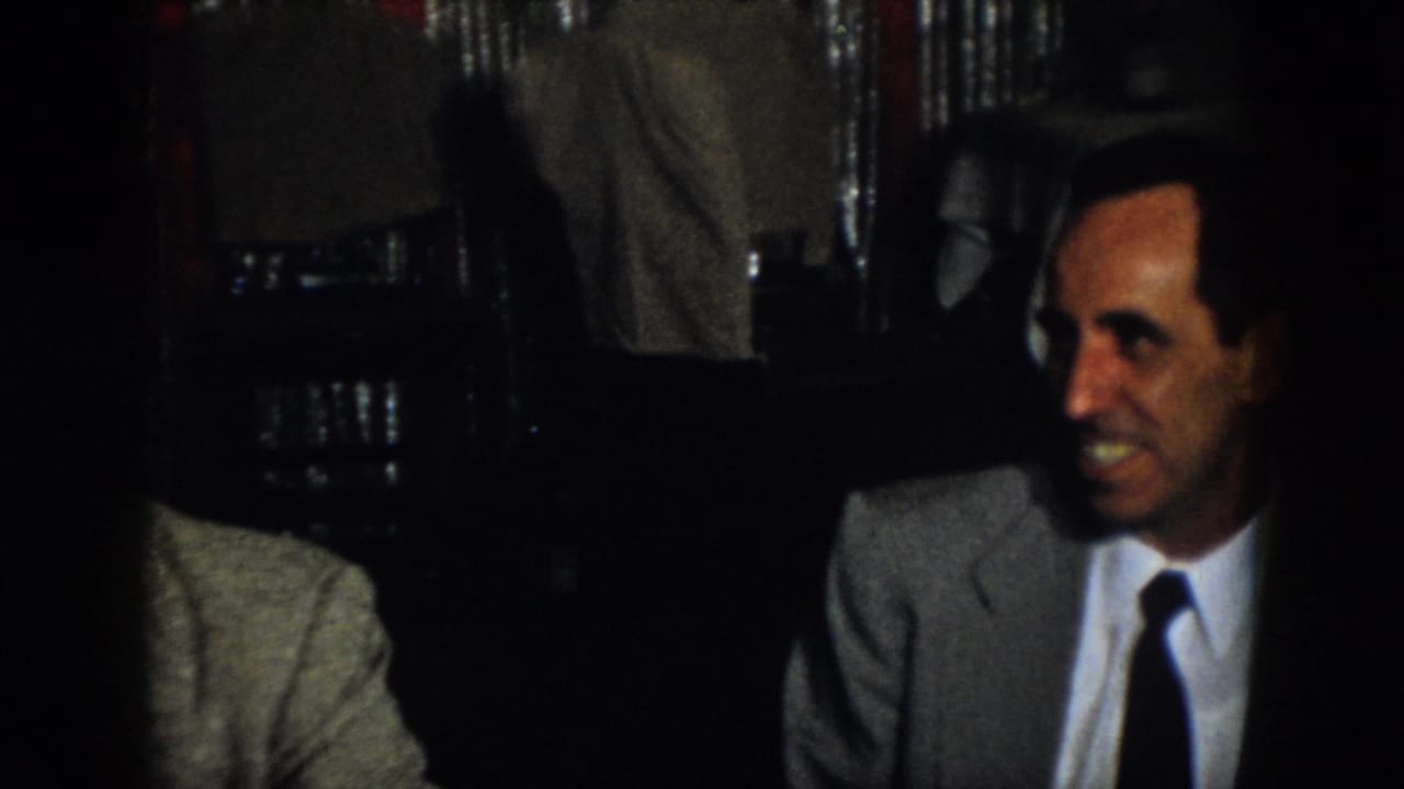 Two adult men in suits sit in a dim interior, with part of another suited person at the left edge.