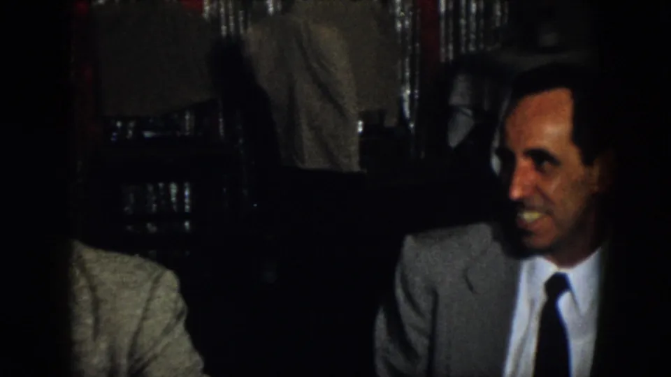 Virginia 1958 home-movie frame showing suited adult men seated indoors in low light during a private social moment.