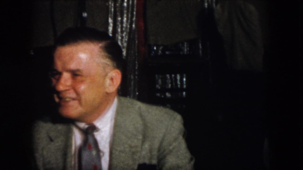 A smiling adult man in a light jacket and tie sits beside another suited man in a dark interior.