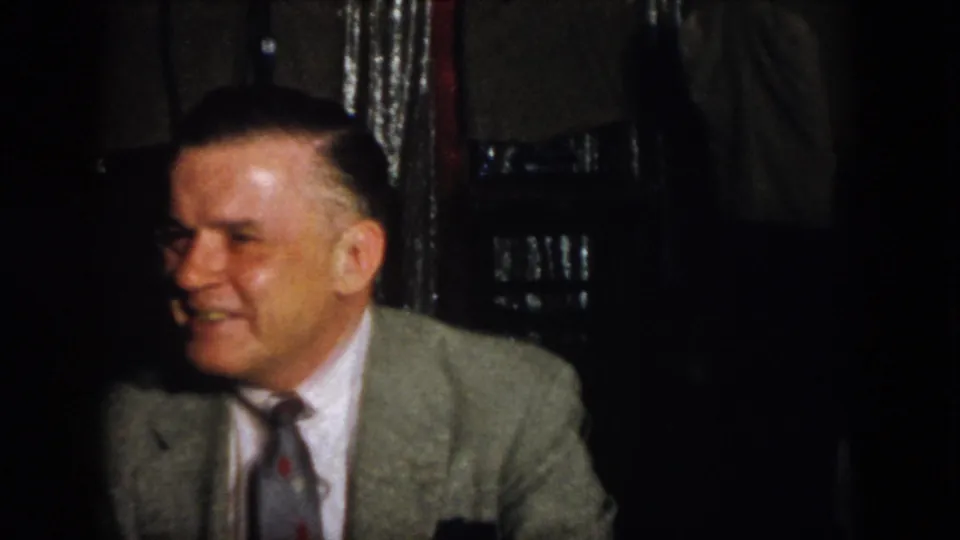 Virginia 1958 frame with a smiling man in a light suit jacket and tie seated in a dim indoor gathering.