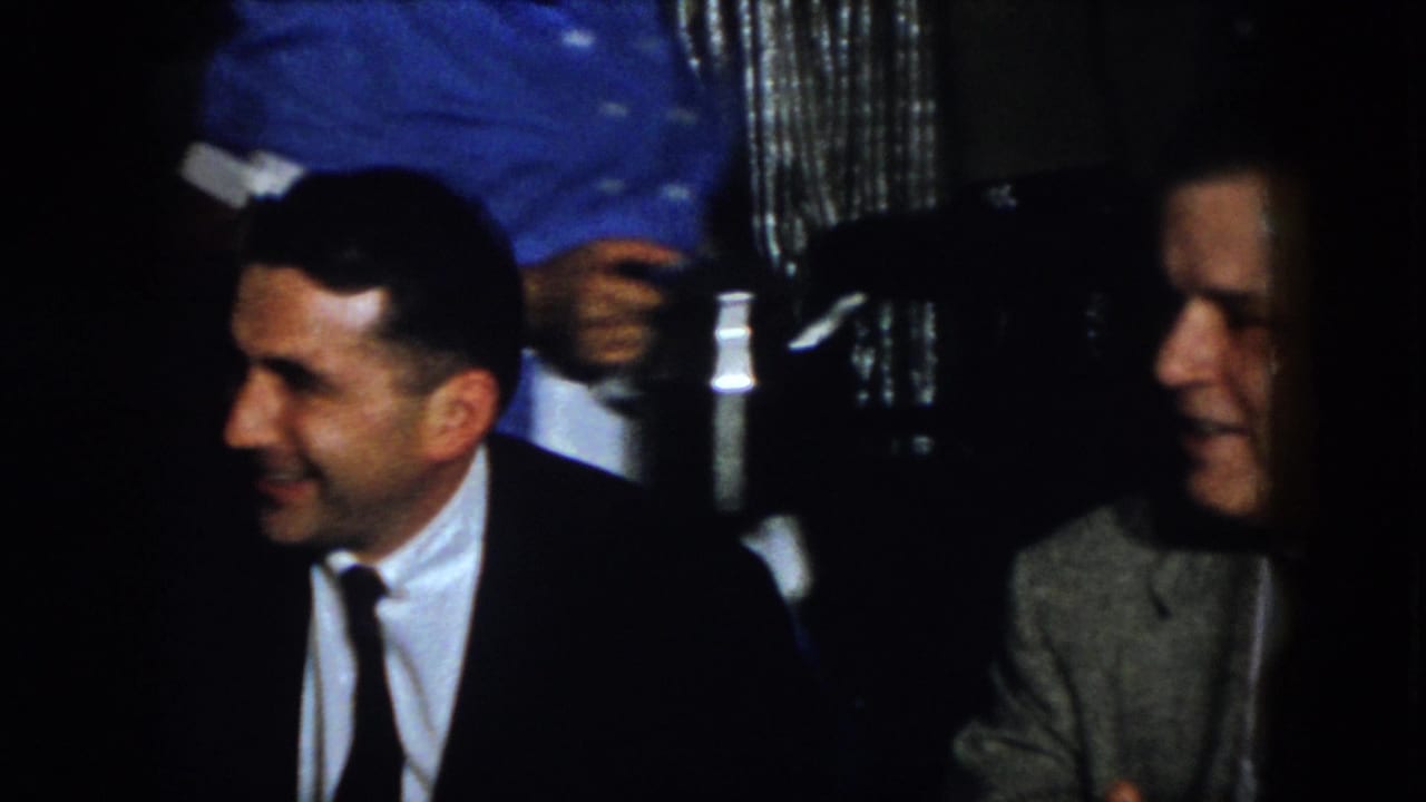 In 1958 Virginia home-movie footage, two suited men sit beside a table with white linens in a dark formal dining interior.