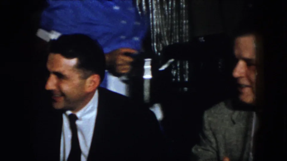 In 1958 Virginia home-movie footage, two suited men sit beside a table with white linens in a dark formal dining interior.