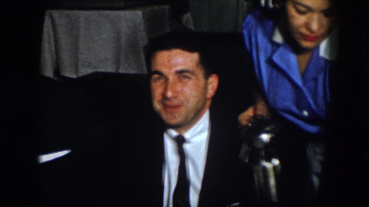 A suited man faces the camera as a woman in a blue uniform pours water nearby.