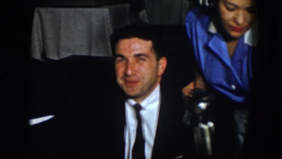 A suited man at the dinner table looks toward the camera while a woman in blue serves water from a metal pitcher.