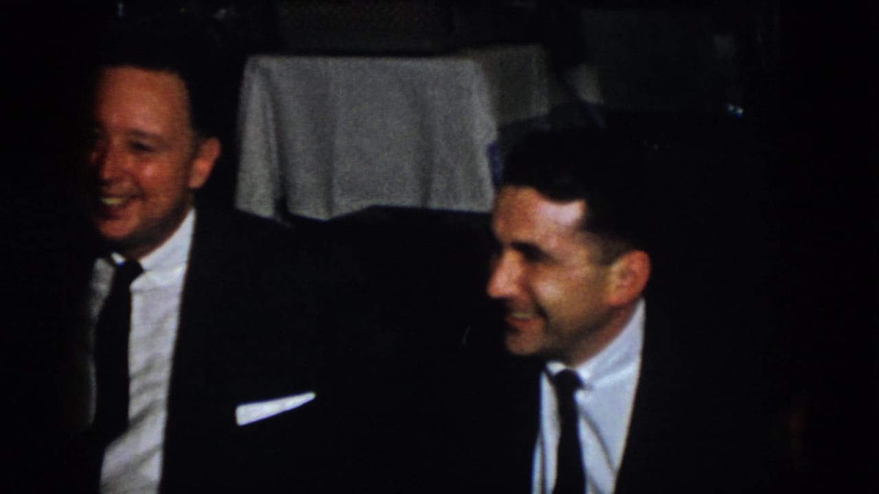 Two adult men in suits smile during conversation at a formal dinner in 1958 Virginia home-movie footage.