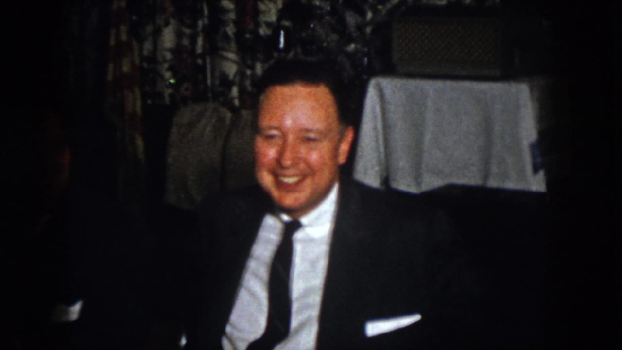 A smiling dinner guest in a dark suit and tie sits near white table linens and patterned curtains in the private interior.