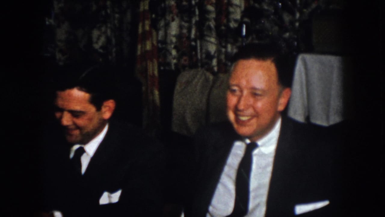 Two suited men at a 1958 Virginia indoor banquet sit close together, one looking down while the other smiles broadly.