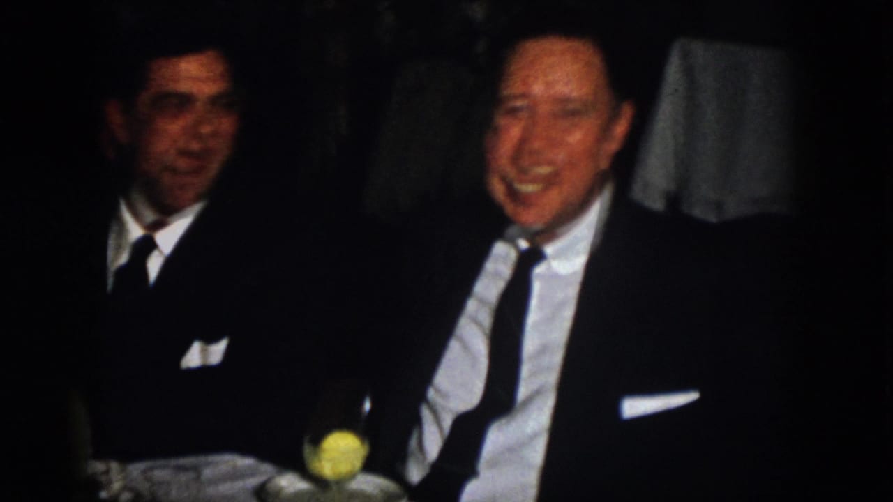 At a dim 1958 Virginia banquet table, two formally dressed men sit near a yellow drink, one smiling toward the camera.