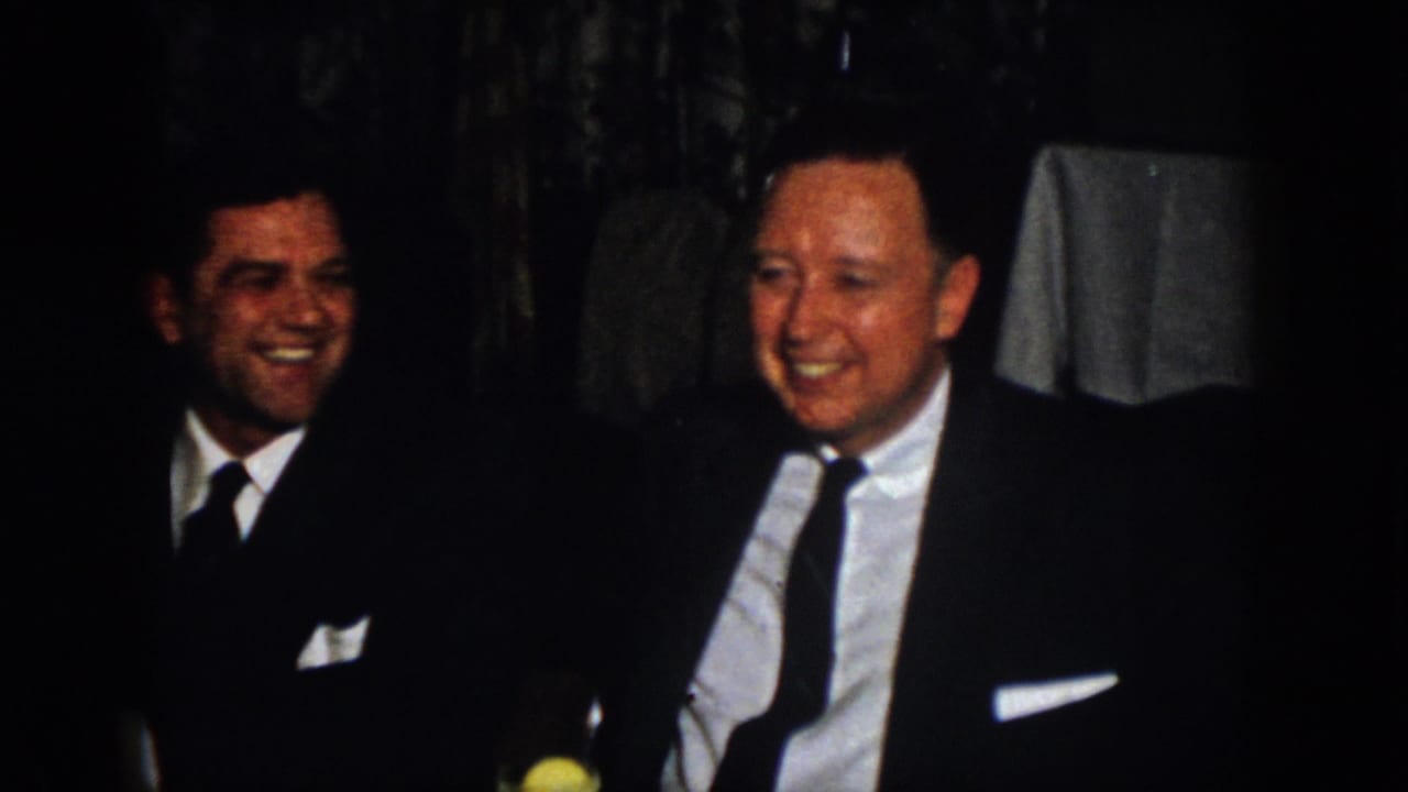 Two men in formal suits laugh together during a 1958 Virginia indoor gathering, with curtains and a yellow drink visible nearby.