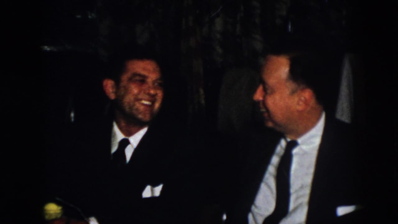 In 1958 Virginia home-movie footage, two suited men sit in a dark interior beside a table setting as one smiles during conversation.