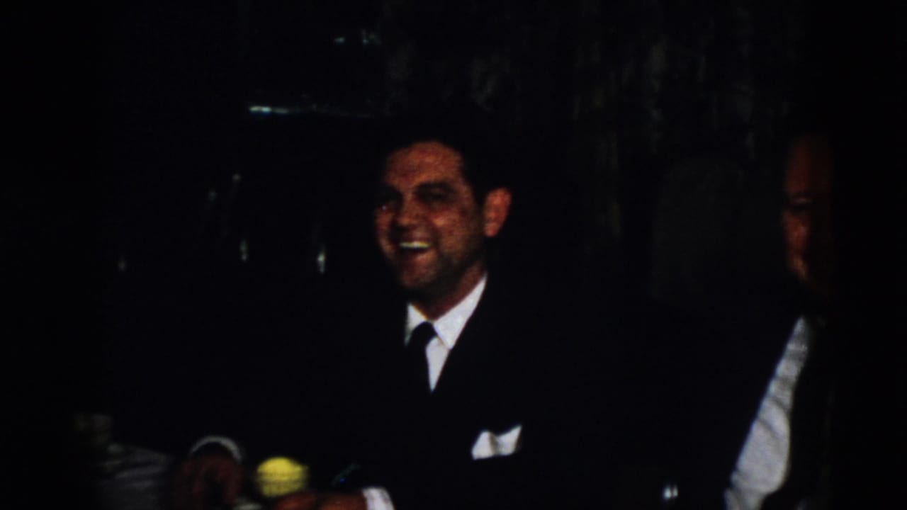 A suited man smiles broadly in the dark 1958 Virginia interior, with part of another seated man visible at the edge of the frame.