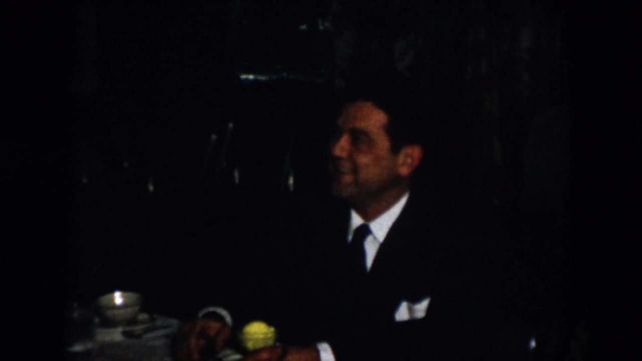 The home-movie frame shows a suited man seated in profile near a teacup and yellow flower in the dim Virginia interior.