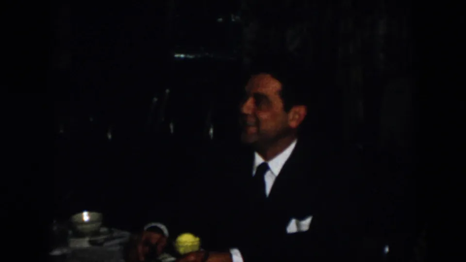 The home-movie frame shows a suited man seated in profile near a teacup and yellow flower in the dim Virginia interior.