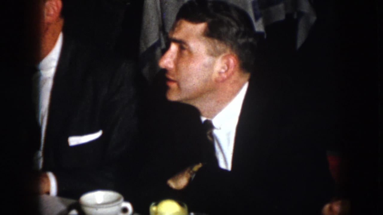 A man in a dark suit and tie is seen in profile beside a white cup, with another suited figure partly visible in the dim 1958 Virginia room.