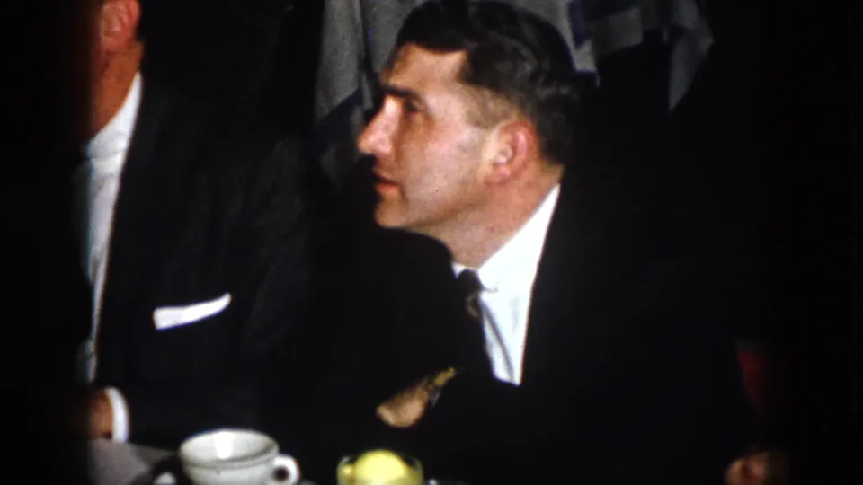 A man in a dark suit and tie is seen in profile beside a white cup, with another suited figure partly visible in the dim 1958 Virginia room.