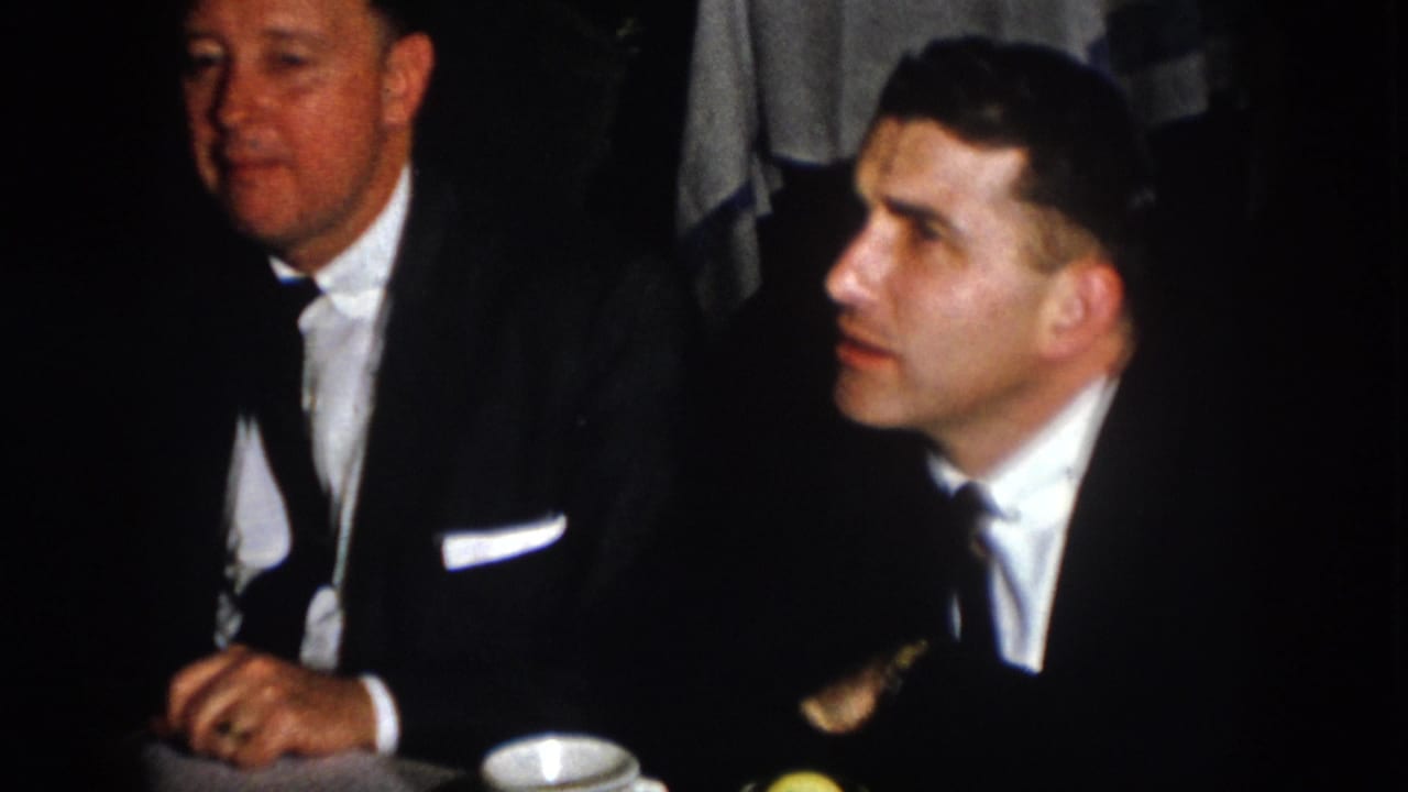 Two adult men in dark suits and ties sit at a Virginia banquet table in 1958 home-movie footage.