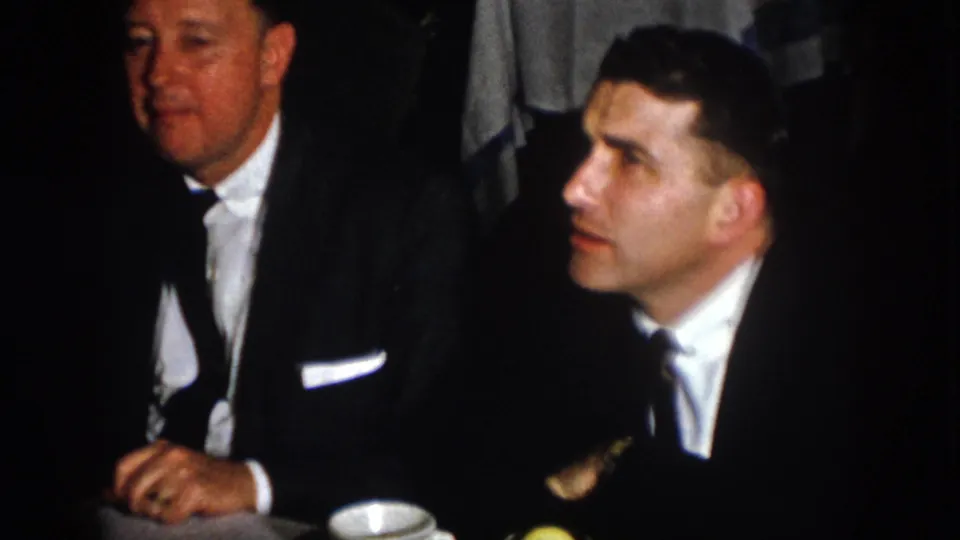 Two adult men in dark suits and ties sit at a Virginia banquet table in 1958 home-movie footage.
