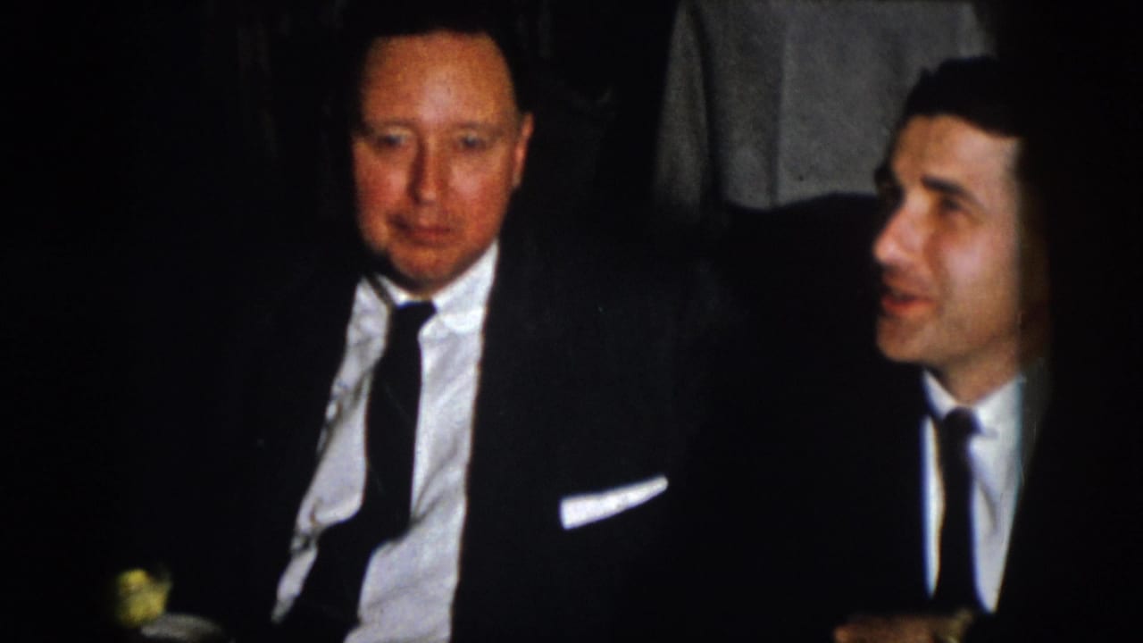 Two formally dressed adult men are framed at a banquet table, with one looking toward the camera area.