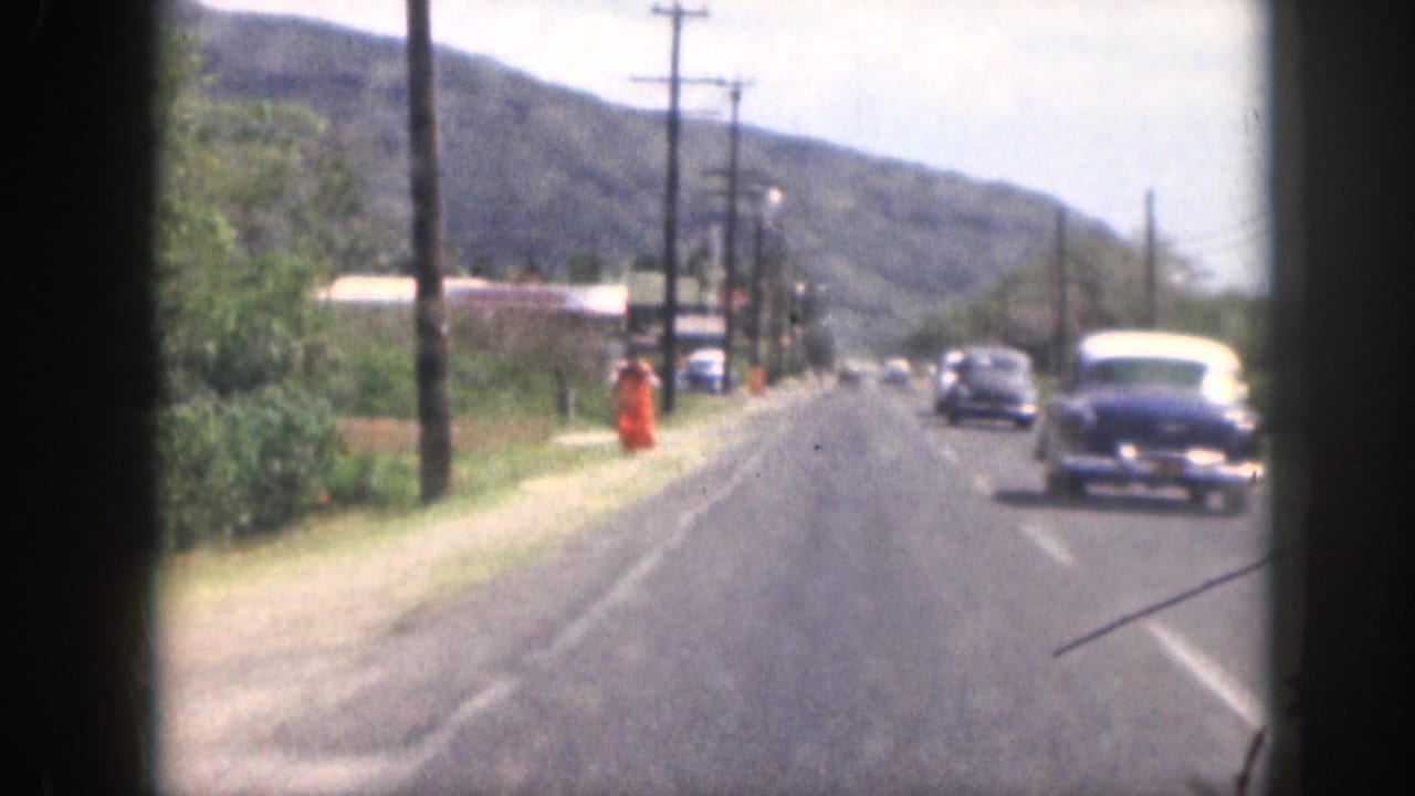 1959 Hawaii home-movie view from a moving vehicle showing vintage cars on a two-lane road with hills, poles, and an orange roadside figure.