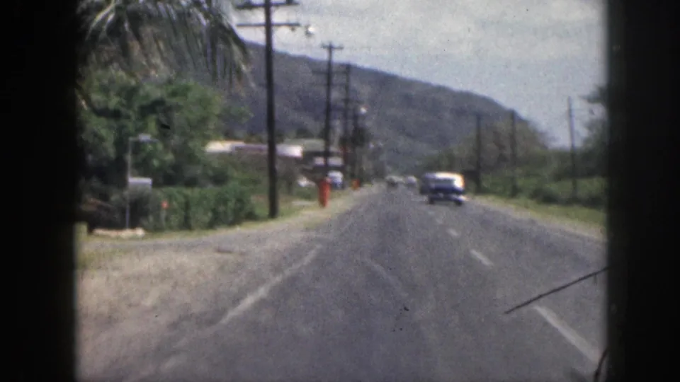 1959 Hawaii road scene from a moving vehicle, with a dark vintage car ahead, roadside poles, green hills, palm foliage, and an orange-clad figure.