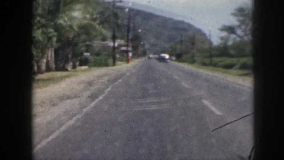 1959 Hawaii travel footage looking down a paved road from a moving vehicle, with distant cars, roadside structures, poles, and green hills.