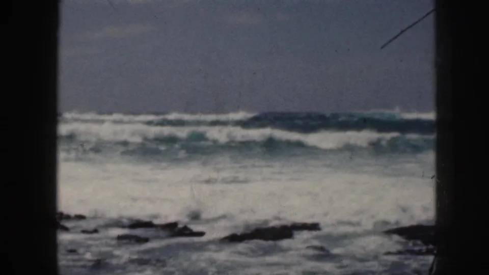 1959 Hawaii home-movie coastal cutaway showing waves breaking across dark rocks and pale water under a hazy sky.