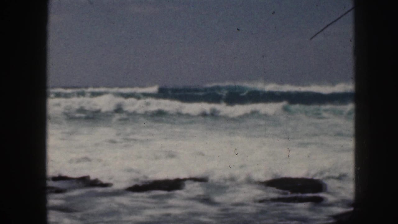 1959 Hawaii 8mm frame showing white surf rolling toward dark shoreline rocks under a muted sky.