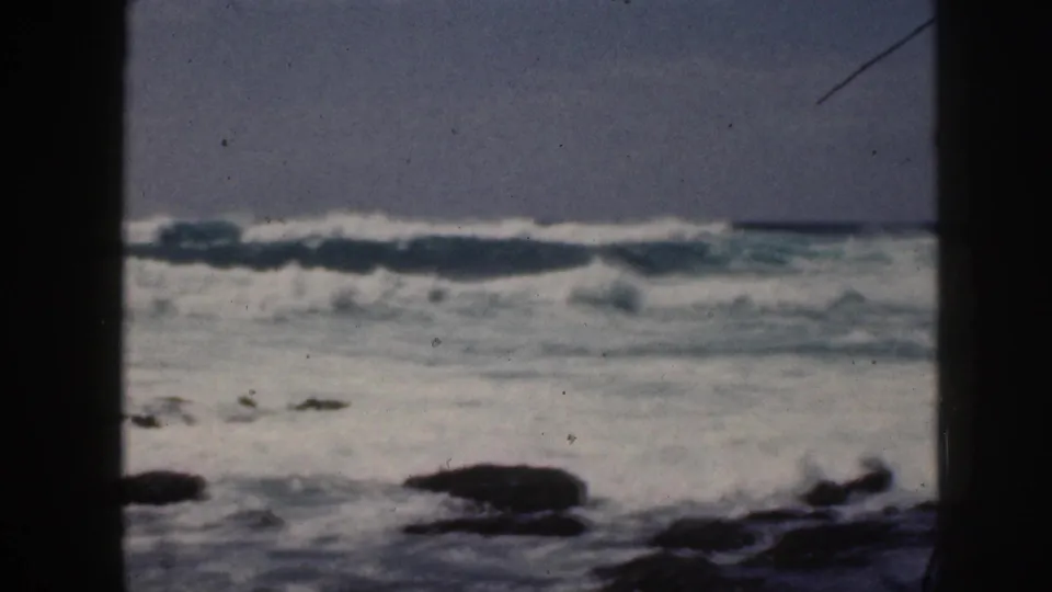 1959 Hawaii 8mm frame with rough surf, dark shoreline rocks, and a central black divider from the film or shooting position.