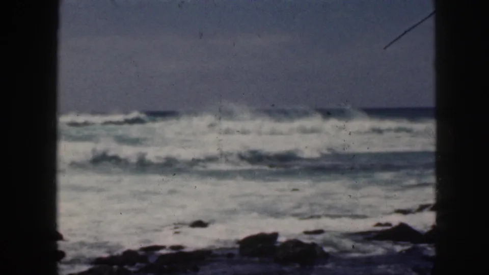 1959 Hawaii 8mm frame of foamy surf crashing near dark rocks, with visible grain, scratches, and shadowed frame edges.