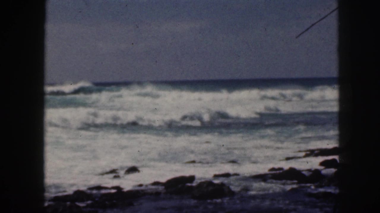 1959 Hawaii home-movie frame of rough surf breaking beyond dark rocks, seen through heavy vertical edge framing.